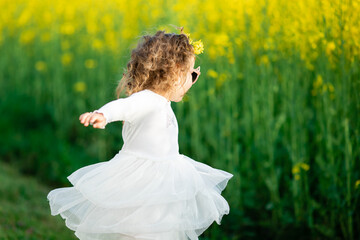  A little girl in a white dress and cat-shaped pink sunglasses stands by a bright yellow flower...