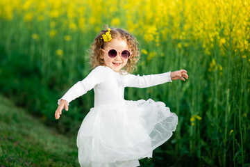  A little girl in a white dress and cat-shaped pink sunglasses stands by a bright yellow flower...