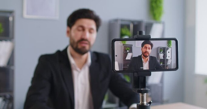 Portrait of a young business man blogger using mobile phone on a tripod recording a video blog from his office. Man employee chatting live webinar using smartphone sitting at workplace. 4k video. - Powered by Adobe