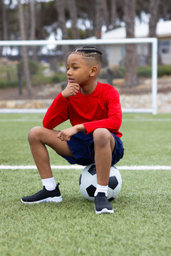 Young boy in red shirt sitting on soccer ball on field, thinking deeply