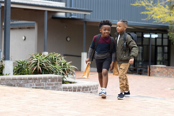 Two diverse boys walking outside school, carrying backpacks and lunch bags, smiling together