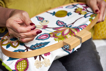 Front close-up view of a woman's hands embroidering a decorative pattern on fabric stretched in an...
