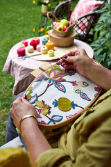 Over-the-shoulder close-up view of a woman embroidering a pattern on fabric held in an embroidery hoop. The hands and hoop are in focus, while her shoulder and back are softly blurred in the foregroun