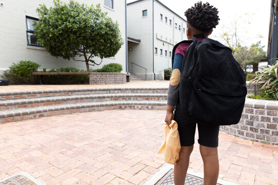 African american boy with backpack walking towards school building, holding lunch bag