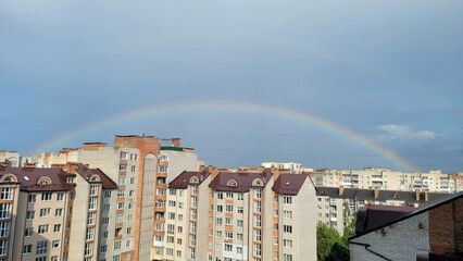 Naklejka premium A vibrant rainbow arches over a cityscape of residential buildings in a serene and picturesque urban landscape after a summer rain, creating a beautiful and hopeful atmosphere.