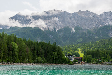 A serene alpine lake with crystal-clear turquoise water mirrors the snow-capped Zugspitze and pine-covered hills. Rowboats glide gently under a clear blue sky—pure alpine tranquility.