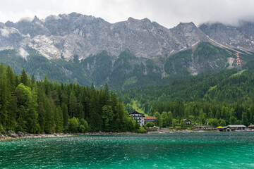 A serene alpine lake with crystal-clear turquoise water mirrors the snow-capped Zugspitze and pine-covered hills. Rowboats glide gently under a clear blue sky—pure alpine tranquility.