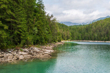 A serene alpine lake with crystal-clear turquoise water mirrors the snow-capped Zugspitze and pine-covered hills. Rowboats glide gently under a clear blue sky—pure alpine tranquility.