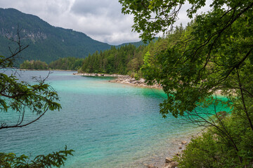 A serene alpine lake with crystal-clear turquoise water mirrors the snow-capped Zugspitze and pine-covered hills. Rowboats glide gently under a clear blue sky—pure alpine tranquility.