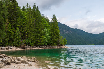 A serene alpine lake with crystal-clear turquoise water mirrors the snow-capped Zugspitze and pine-covered hills. Rowboats glide gently under a clear blue sky—pure alpine tranquility.