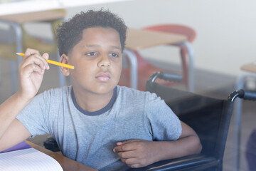 In classroom, boy in wheelchair holding pencil, thinking deeply about lesson, at school
