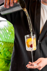 A bartender pours a refreshing cocktail from a metal shaker into a wine glass filled with ice and...