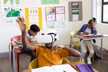 In classroom, african american boy using VR headset while others focus on writing tasks, at school