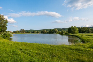 Fototapeta premium Freshwater fishing pond in the countryside with grassy banks and forested hills. Used for aquaculture and angling of carp and other freshwater fish species.