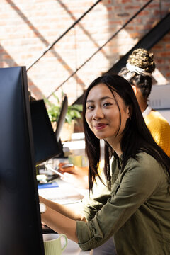 Asian woman coding on computer in modern office, smiling confidently