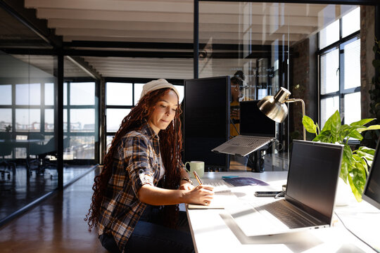 Woman coding at desk in modern office, writing notes and focusing on work