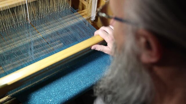 Weaver at the traditional loom. Mature man with a graying beard and glasses using a weaving shuttle works at the loom, weaving cloth and tapestry by hand in textile garment production, slow motion