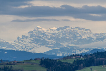 Majestic snow-capped mountains rise above lush green valleys under a cloudy sky in early spring