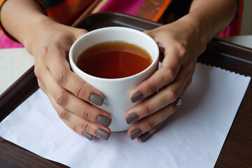Close up of woman's hands with grey manicure holding white mug of hot tea on tray, enjoying relaxing moment during calming afternoon break in cafe or at home