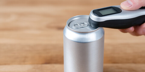 A silver can is placed on a wooden surface, and a person holds a digital thermometer to measure its temperature. Beverage temperature measurement 