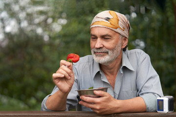 Fototapeta premium The health benefits of strawberries. Senior citezen on a garden terrace with a ripe berry in one hand and a bowl of berries in the other hand against a blurred natural background.