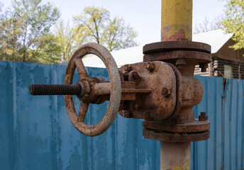Old rusty industrial valve on a yellow metal pipe near a blue fence outdoors