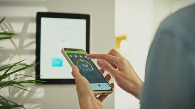 A person controls the home temperature using a smartphone app, interacting with a smart display on the wall. This takes place in a bright, contemporary living room