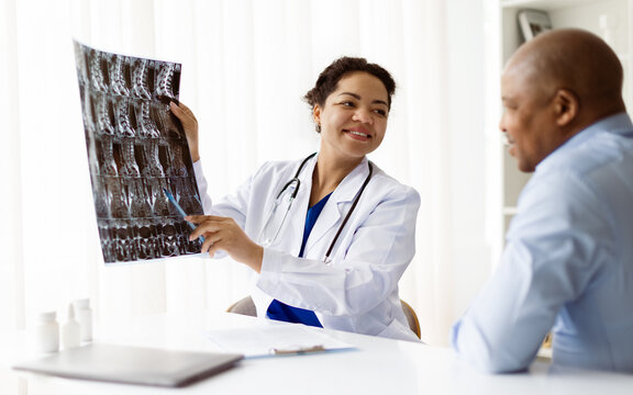 Smiling Black Doctor Woman Showing X-Ray To Male Patient During Appointment In Clinic, Therapist Lady In Uniform Consulting Man During Meeting In Hospital, Discussing Treatment