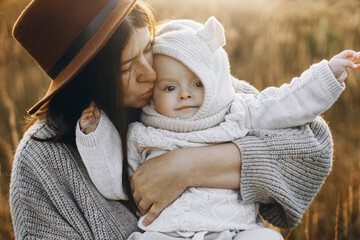 Happy mother hugging and kissing cute baby son in autumn field in soft evening light. Parenthood. Mom and little child in cozy stylish clothes embracing in fall park, love atmospheric moment