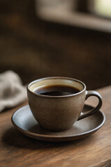 Obraz premium A warm cup of dark coffee sits on a saucer beside a linen napkin on a rustic wooden table, creating a cozy and inviting atmosphere.