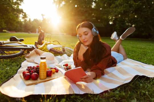 Carefree woman listening to music in headphones, lying on picnic mat and reading paper book in green park on summer sunny day - Powered by Adobe