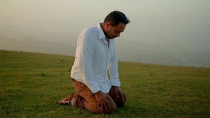 Indian man kneeling in prayer on a green field, wearing a white shirt.  

