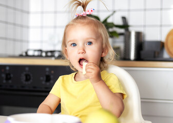 Adorable toddler girl with blond hair and bow eating healthy snack in kitchen while sitting in high chair. Concept of child nutrition, development and family care