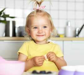 Cute blonde toddler girl in yellow shirt smiling while eating healthy breakfast in kitchen. Concept of happy childhood, family care and healthy nutrition