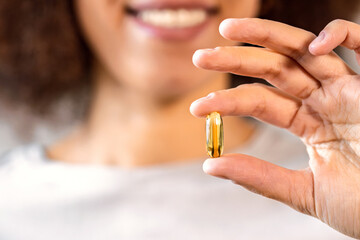 Close up of female hand holding golden omega 3 capsule with blurred smiling face in background. Concept of health supplement, nutrition and self care