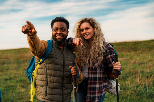 Happy diverse couple hiking in a grassy field, man pointing at something in the distance. They are wearing casual outdoor clothing and backpacks, enjoying a sunny autumn day.