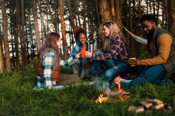 Friends gathering for drinks around woodland campfire. Authentic moment showing diverse group sharing thermos drinks during sunset camping adventure.