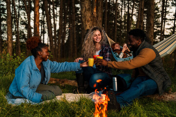 Friends relaxing by campfire in woodland setting. Authentic outdoor lifestyle capture showing...