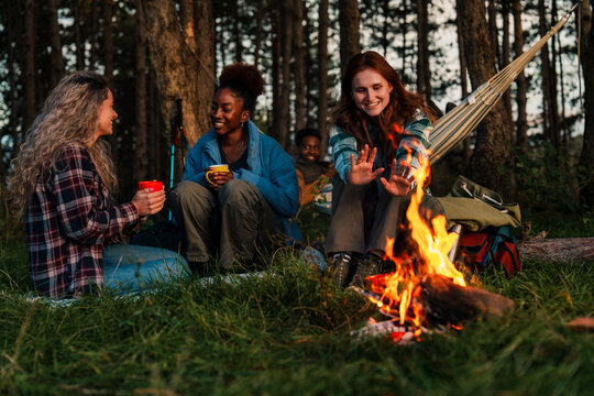Golden hour camping with friends warming up by fire. Genuine laughter and cozy atmosphere as diverse group enjoys evening drinks in forest setting. - Powered by Adobe