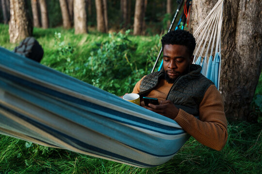Young man relaxing in hammock while checking phone during forest camping. Modern outdoor lifestyle blending nature and connectivity in peaceful setting. - Powered by Adobe