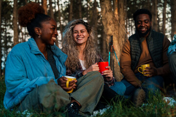 Sunset camping scene with friends sharing stories over colorful mugs. Natural lifestyle photo showing genuine smiles and diverse outdoor connections.