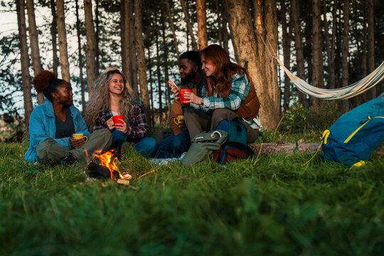 Friends creating cozy camping atmosphere in woodland setting. Authentic moment capturing diverse group sharing stories and drinks at golden hour.