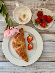 Cup of coffee, croissant and strawberries on the white plates and fresh peony flower on the wooden background