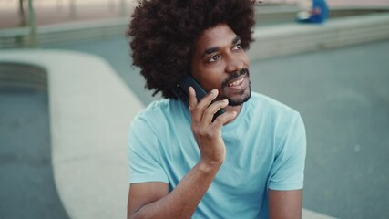 Closeup young African American man in light blue t-shirt sitting on city park bench and talking on smartphone. Camera moving forwards approaching to the person. lifestyle concept. slow motion - Powered by Adobe