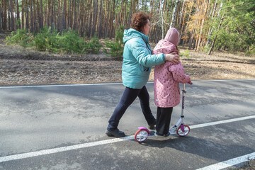Little girl riding scooter while walking with her grandmother in the forest on a sunny day
