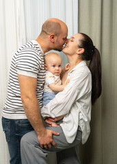 Fototapeta premium Loving mom and dad hold their one year old son in their arms and kiss gently. Warm and sincere moment reflecting family love, care and closeness.