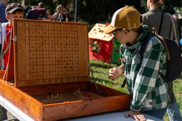 Curious boy in checkered shirt and cap playing large wooden battleship game outdoors on sunny day during family festival. Concept of childhood fun, outdoor games, learning through play and strategy.
