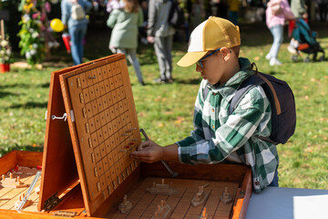 Boy wearing plaid shirt and baseball cap concentrates on playing giant wooden board game outdoors during public event. Concept of educational entertainment, strategic thinking, and childhood joy.
