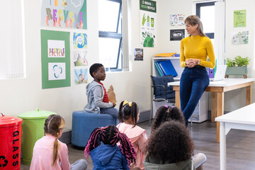 Female teacher engaging diverse students in classroom recycling lesson, kids listening, at school