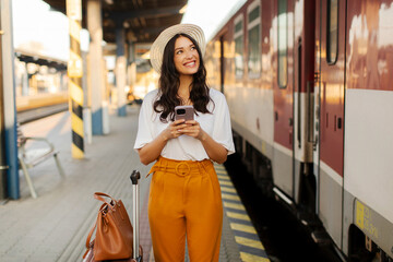 Happy woman tourist using cellphone and looking at wagon number, waiting for boarding on train, standing at railway station platform
